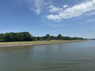 This is a wide-angle, eye-level shot of a calm, grey waterway under a bright blue sky with scattered white clouds. The water fills the foreground and midground, showing gentle ripples. The background features a lush, green treeline along the far bank, interspersed with some buildings and parked vehicles. A paved walkway runs along the edge of the water, bordered by a stone embankment and then a grassy slope leading up to the trees. A solitary white pole stands on the embankment, and a dark bench is visible on the grass. Further along the bank, the scene curves out of view, revealing more distant structures and what appears to be a harbor. The overall impression is one of a serene, possibly industrial or port area on a clear, sunny day, likely late morning or early afternoon given the bright light. There is no visible text in the image.