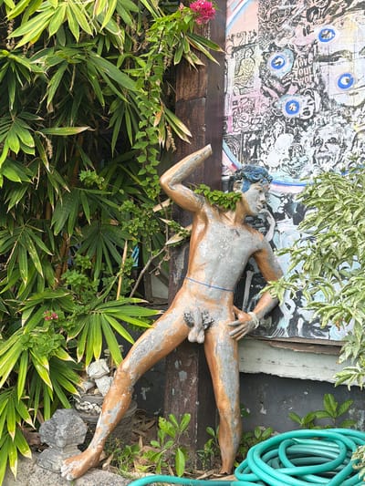 In Tejakula, Indonesia, a life-sized statue of a male figure stands in a garden setting. The statue, with metallic gold and silver coloring, is partially covered by green vines. It depicts a nude male with blue-painted hair, one arm raised and bent, and the other resting on his hip. The statue is positioned next to a wooden post, with a vibrant, abstract mural visible on a wall behind it. Lush green bamboo foliage surrounds the left side of the scene, and a coiled green garden hose lies on the ground in the foreground to the right. Small, grey stone decorative elements are scattered around the base of the statue.