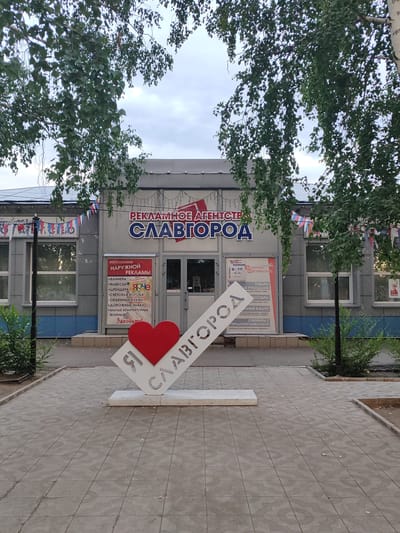 This media file captures the exterior of a building in Slavgorod, Russia, likely a business. The building's facade is a light gray with blue accents near the bottom. Above the entrance, prominent Russian text reads "РЕКЛАМНОЕ АГЕНТСТВО СЛАВГОРОД" which translates to "ADVERTISING AGENCY SLAVGOROD."
In front of the building, a prominent white sculpture in the shape of an angled "I" followed by a heart and the word "СЛАВГОРОД" (Slavgorod) is displayed. This suggests a sense of local pride or a tourist attraction. The sculpture is made of white material with cut-out letters.
A red heart is positioned on the "I" part of the sculpture, indicating affection or love for the city. The building features multiple windows and two glass doors for entry. To the left of the main entrance, a smaller red sign lists services offered by the agency, including banners, signs, light boxes, volumetric letters, road signs, and small architectural forms, with the word "Заходите!" (Come in!) at the bottom. To the right, another sign indicates "Багетная Мастерская" (Framing Workshop) and mentions "ТОВАРЫ ДЛЯ РУКОДЕЛИЯ" (Craft Supplies) and "СУВЕНИРЫ С ГОРОДСКОЙ СИМВОЛИКОЙ" (Souvenirs with city symbols).
The scene is framed by the branches of leafy trees, suggesting it's a daytime shot with overcast or cloudy skies. The ground in front of the building is paved with square tiles. Small, colorful Russian flags are strung along the building, adding a festive touch. The overall impression is a commercial establishment with a welcoming and community-oriented message.