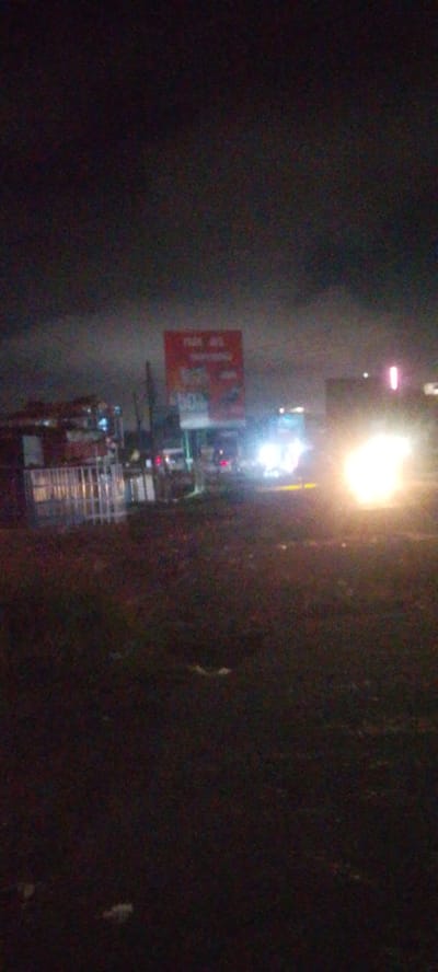 The image depicts a nighttime scene in Ndenderu ward, Kenya, characterized by low light and artificial illumination. The sky is dark and appears overcast.
In the foreground, the ground is dark, uneven, and appears unpaved, possibly dirt or gravel, with some faint light-colored debris scattered across it.
The midground features a roadside environment. To the left, a cluster of indistinct structures, possibly commercial stalls or buildings, are visible, dimly lit from within or nearby. In front of these structures, a white, slatted fence or barrier runs horizontally. In the center-right, a prominent rectangular billboard with a red background and white text is erected. Partially legible text includes "FARA HA" at the top and "10%" further down.
A road runs across the frame, with visible vehicle lights. On the far right, the intense, bright white headlights of an approaching vehicle create significant glare. Further to the left of these headlights, a vehicle with bluish-white lights is also discernible. Red tail lights or brake lights are visible further back and to the left on the road. A faint, vertical red light is present on the far right edge of the frame, possibly part of a sign or traffic light.
No individuals are clearly visible or identifiable in the image. The scene captures a moment along a busy-looking road at night, characterized by artificial lights against a dark, semi-urban backdrop.