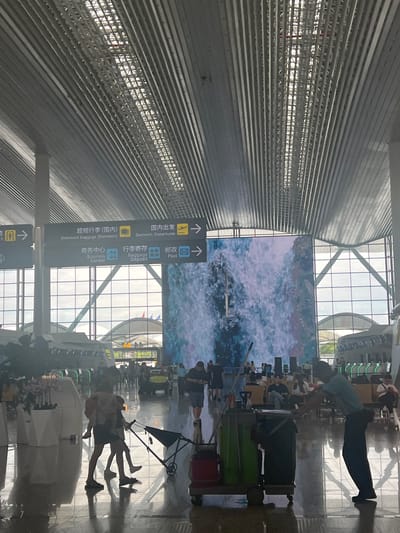 This image depicts the interior of a modern airport terminal, likely in Guangzhou City, China, given the bilingual signage. The architecture is characterized by a high, ribbed metal ceiling and large glass windows that allow natural light to illuminate the space. A massive digital screen displaying a waterfall is a prominent feature.
In the foreground, a person, appearing to be a cleaner, is attending to a cleaning cart equipped with buckets and supplies. To the left of the cleaner, a woman is carrying a child on her back while pulling a stroller. Several other individuals and groups of people are visible throughout the terminal, some seated in waiting areas and others walking. Airport infrastructure, such as signage indicating "Oversized Baggage (Domestic)", "Domestic Departures", "Business Center", "Baggage Deposit", and "Post", is clearly visible. The floor is highly reflective, mirroring the ceiling and figures. In the background, airplane fuselage-like structures are partially visible, suggesting the proximity to departure gates or a maintenance area. The overall scene conveys the busy atmosphere of a transportation hub.