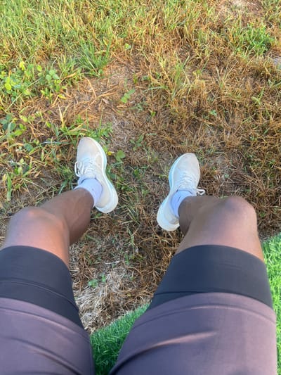 The image shows a first-person perspective of a person sitting on dry grass in Dam Camp, Nigeria. The person is wearing white athletic shoes and dark athletic shorts. Their legs and knees are visible. The background consists of dry, brown grass mixed with some green patches and small weeds. The lighting suggests it is daytime.