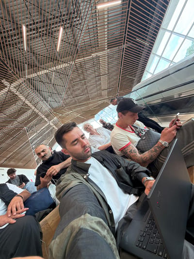 The image depicts a group of people sitting in what appears to be an airport or train station waiting area. The architecture features a distinctive wooden slatted ceiling and a large window letting in natural light. In the foreground, a man in a grey and black jacket is holding a black laptop with a "ThinkPad" logo. To his right, another man wearing a baseball cap and a white t-shirt with red trim is looking at his phone. Behind them, several other individuals are seated, some also looking at their phones or resting. The overall impression is one of people waiting, possibly for travel, in a modern, well-lit interior space. There is no specific indication of the location being in Ianeti, Georgia.