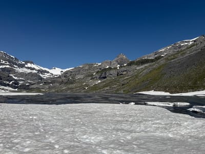 The image depicts a mountain landscape in Leukerbad, Switzerland, under a clear blue sky. In the foreground, a large expanse of textured snow covers the ground, appearing somewhat melted and uneven. Beyond the snow, a dark, rocky surface with patches of water is visible, suggesting a partially frozen lake or riverbed. Further in the background, imposing mountains rise, characterized by jagged gray rock faces interspersed with patches of white snow. The slopes of the mountains are partially covered with green vegetation. The overall scene conveys a sense of rugged, alpine wilderness during a period of transition, possibly late spring or early summer, when snow is melting. There are no people or animals visible in the image.