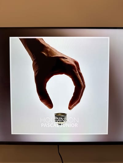 This media file features a close-up of a person's hand, silhouetted against a bright, white background, holding a small, cylindrical object. The hand is positioned in a way that suggests it is carefully grasping or presenting the object, with the fingers curved around it. The object itself appears to be metallic with a ridged texture.

To the right of the hand and object, there is text printed vertically. The visible words are "HOLDING" and "PASCAL JUNIOR". The overall presentation is clean and minimalist, with the focus primarily on the hand and the object it holds.

The setting appears to be an indoor environment, possibly a studio or a gallery, given the controlled lighting and the nature of the displayed image which looks like a print or a digital display. There are no external environmental cues like time of day or weather. The emotional tone conveyed is one of focus and precision, highlighting the act of holding.

The context provided indicates this is from Khan Mean Chey, Cambodia.