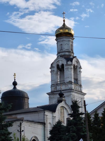 This is a low-angle, outdoor, eye-level shot of the Church of the Assumption of the Blessed Virgin Mary in Rogachovo, Russia. The church features a tall, white bell tower with a golden dome, topped with a cross. To the left of the bell tower is a darker dome, also topped with a cross. The sky is a bright blue with scattered white clouds. A dark wire stretches across the upper portion of the frame. Trees, some of which appear to be fir trees, are visible in the foreground and partially obscure the lower parts of the church building. The church appears to be made of white stone and has a long, dark roofline. The overall scene depicts a religious building against a cloudy sky.