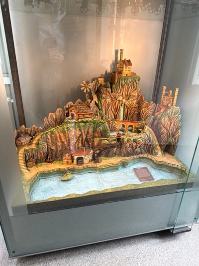 The image shows a three-dimensional, intricately detailed model of a landscape with buildings, mountains, and a river. The model is housed inside a glass display case.

Buildings are situated on the mountains, including structures resembling castles and smaller houses. A windmill is also present. A bridge connects sections of the landscape. The river is rendered in light blue.

A sign is affixed to the river's edge, likely providing information about the model.

The scene is indoors, with the model presented as an exhibit. The glass display case is modern, with a dark gray frame.

The location is Biberach an der Riß, Germany.