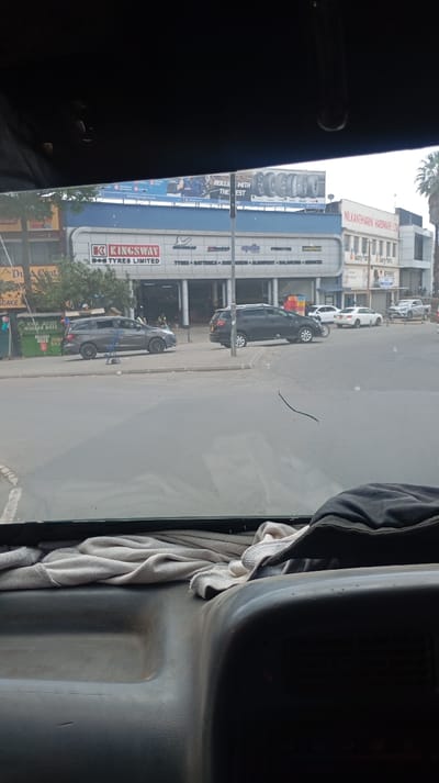 The image is a first-person perspective shot taken from inside a vehicle, looking out through the windshield at an urban street scene. The top and bottom portions of the image show the vehicle's interior, specifically the visor/ceiling and the dashboard, respectively.

In the foreground, the dashboard of the vehicle is visible, dark-colored and somewhat curved. On top of the dashboard, a light-colored, possibly white or off-white, cloth or towel is crumpled. To the right of the cloth, a dark jacket or piece of clothing is also resting on the dashboard. The windshield has a visible, somewhat serpentine crack or scratch running horizontally across its lower-middle section.

The midground features a paved road with a curb and sidewalk on the right. Several vehicles are parked or driving. To the left, a dark grey or black minivan-like vehicle is parked on the sidewalk near a green kiosk. Further right and slightly behind it, a dark-colored SUV is also parked partially on the sidewalk or near the curb. Other cars, including a white sedan and a silver sedan, are visible further down the street on the right.

The background is dominated by commercial buildings. The most prominent building, centrally located, is a tire shop. Its facade is light blue and white, with large windows. Above the entrance, a large sign reads "K KINGSWAY TYRES LIMITED." Below this, smaller signs list brands and services: "MICHELIN," "BFGoodrich," "APOLLO," "Pirelli," "TYRES * BATTERIES * SUSPENSION * ALIGNMENT * BALANCING * SERVICES." To the far left of this building, a small green kiosk has "MPESA" visible on it, along with text "MR JULIUS MUGORA BAZE" and "NYUMBA KWA..." partially readable. Another sign next to it reads "DUA COAT."

Above the Kingsway Tyres building, a large billboard is visible, advertising tires. Part of the text reads "ROLL WITH THE EST" (likely "ROLL WITH THE BEST") and shows various tires.

To the right of the Kingsway Tyres building, another multi-story building is visible with a sign that reads "NILKANTHVARN HARDWARE LTD." Below this, another sign reads "Glory Har." A palm tree is visible on the far right, near this building.

The sky appears overcast but bright, suggesting daytime. The overall impression is a typical busy commercial street.