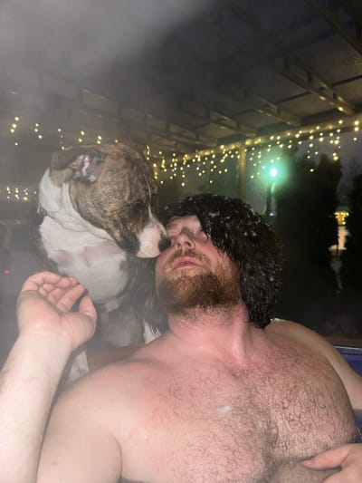 The image depicts an outdoor night scene in the city of Базарово, Russia, featuring a male and a dog in steaming water. A Caucasian male, viewed from the chest up, is partially submerged in a hot tub or similar heated pool. He possesses dark, wet hair with white specks consistent with snow or ice, and a reddish-brown beard. His eyes are partially closed, and his mouth is slightly agape. His left arm is raised with the hand visible. Resting its upper body near the man's left shoulder, a brindle and white dog, consistent with an American Staffordshire Terrier or similar bully breed, is present. Its muzzle is in contact with the man's upper lip/nose area. Overhead, a wooden structure, possibly a pergola or gazebo, is adorned with warm yellow string lights. Visible steam rises from the water. In the dark background, a distant artificial light source emits a greenish hue, and faint outlines of trees are discernible.