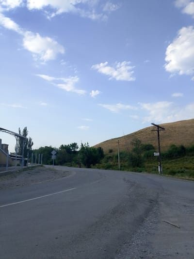The image depicts a rural roadside scene in Temen-Su, Kyrgyzstan. A paved road curves to the right, with a white line marking its center. On the left, a building with a covered, open-air structure is visible, supported by metal poles, adjacent to a dirt and gravel area. Beyond the building, a line of tall, slender trees stands. Further along the road, a traffic sign with a diamond shape and a horizontal bar is posted. The right side of the image features a grassy, sloping hillside. A wooden utility pole with multiple crossbars, adorned with birds, stands on the hillside, and a smaller utility pole is visible near the road. Several trees and bushes are scattered along the base of the hill. The sky is a light blue with scattered white clouds.