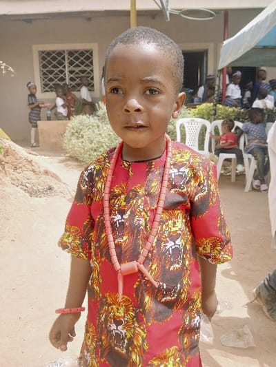 This image depicts an outdoor scene in Masvingo, Zimbabwe, featuring a young Black child as the primary subject, standing centrally and facing the camera. The child wears a short-sleeved, red garment patterned with roaring lion heads in shades of brown and black. A long, salmon-colored beaded necklace with a distinctive pendant hangs from the child's neck, complemented by a matching beaded bracelet on the left wrist. The child's left hand grasps a crumpled clear plastic object.

The ground consists of light brown, loose earth or sand. In the background, a light-colored building with a gridded window is visible on the left. A small dirt mound is present in the mid-ground to the left. Several other children are scattered throughout the background; some are standing near the building, one holding a tray, while others are seated on white plastic chairs. A light blue canopy or tarp is partially visible in the upper right, and a yellow pole extends vertically in the upper left background. Bright daylight illuminates the scene, casting shadows. An adult's leg and foot are partially visible on the far right edge of the frame.