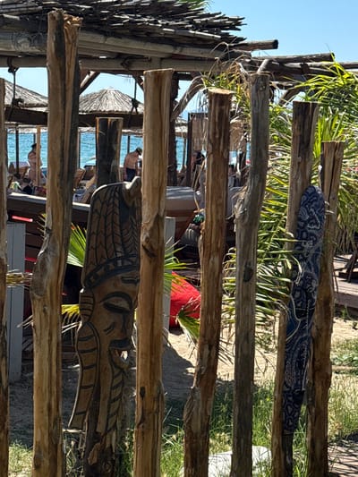 The image depicts a beachside scene in Asprovalta, Greece, focusing on a rustic wooden fence in the foreground. The fence is constructed from several vertical logs, some of which are adorned with carvings. A prominent carved wooden mask, featuring a headdress and intricate facial details, is attached to one of the logs. To the right, a patterned blue fabric is draped over another log, resembling a sarong or towel.
Behind the fence, the scene opens up to a beach setting. Several thatched umbrellas provide shade, and lounge chairs are visible. The sea, with its clear blue water, is in the background. People are present on the beach, some appearing to be relaxing on chairs, while others are standing or walking near the water. One individual, a shirtless man, is seen near the shore. The overall atmosphere suggests a warm, sunny day at a coastal resort. The wooden structure with its thatched roof above the beach area indicates a beachfront bar or restaurant. Green foliage, likely palm leaves, are interspersed throughout the scene, adding to the tropical ambiance.