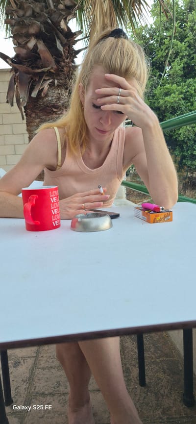 The image shows a woman sitting at a white table outdoors, with a palm tree and foliage in the background. She has blonde hair pulled back and is wearing a light peach tank top and has her hand on her forehead. She is holding a lit cigarette in her right hand. On the table in front of her are a red mug with the word "LOVE" repeated on it, a silver ashtray, a smartphone, a pack of Chesterfield cigarettes, and a pink lighter. The scene appears to be in Almáchar, Spain.
