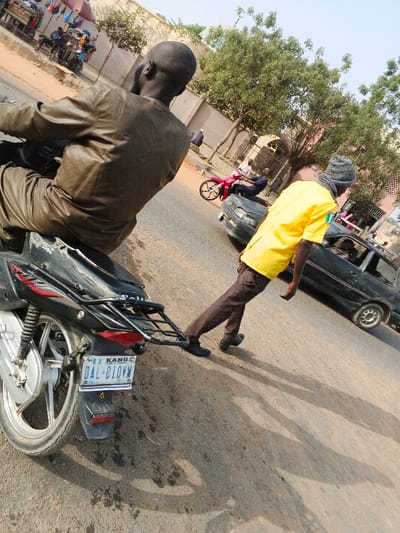 The image displays a street scene in Gobirawa, Nigeria, under clear, sunny conditions. In the immediate foreground, a dark-skinned male with a bald head and beard, dressed in a brown full-length garment, is seated on a black motorcycle. The motorcycle's rear fender features a black cargo rack and a visible license plate reading "KANO DAL-810WM". To the right of the motorcycle, a dark-skinned male wearing a yellow short-sleeved shirt, dark brown cargo pants, black shoes, and a grey knitted beanie is walking forward, his left foot extended.

In the mid-ground, a dark-colored sedan is partially visible, moving from right to left across the frame. Further in the background, a red motorcycle with two occupants is visible. The environment includes light-colored buildings with arched openings, green deciduous trees lining the street, and on the far left, market stalls with colorful canopies and various goods. Long shadows are cast by individuals and objects onto the asphalt and dirt road surfaces.