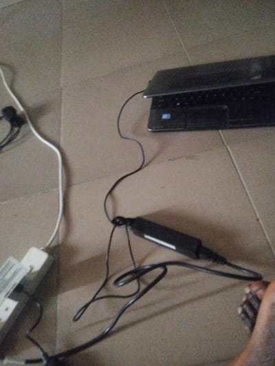 The scene depicts an indoor setting in Kaninkon, Nigeria, with electronic equipment laid out on a tiled floor. A dark grey laptop, partially open with its screen facing away, is positioned in the upper right section. A black power cable connects the laptop to a black power adapter, which features a white label and rests centrally on the light-colored tiled floor. This adapter is further connected by a black cable to a white power strip, partially visible in the mid-left. A white cable, likely an extension cord, extends from the power strip. A bare, dark-skinned human foot is partially visible in the lower right corner, positioned near the laptop's power adapter.