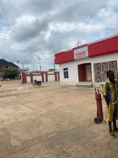 The image shows the exterior of a building labeled "MUNSIR COOKING GAS" in Nassarawa, Nigeria. The building is white with a red trim. In the foreground, a person wearing a colorful striped shirt and shorts stands next to a red gas cylinder on wheels. A motorcycle is parked in the middle ground. The area is paved with concrete, and there are white metal barriers. In the background, a building under construction is visible, along with trees and power lines. The sky is cloudy.