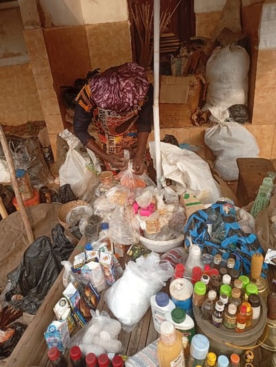 The image depicts a vibrant and cluttered market stall in Itu, Nigeria, focusing on a person actively engaged in preparing goods for sale. The central subject, likely a woman, is seen from a slightly elevated angle, bending over a heap of merchandise. She wears a patterned head covering, possibly purple and white, and a colorful garment over dark long sleeves. Partial text, including "FREE INT..." and "INTERNATIONA[L]," is visible on her clothing. Her hands are busy tying a clear plastic bag containing orange-hued items, suggesting packaging or portioning.

The setting is an densely packed market stall, with merchandise spilling across a wooden surface that appears to be a counter or floor. The background features light brown tiled walls, indicating a fixed structure. Against these walls, large white sacks filled with goods, cardboard boxes, and various bundles are stacked. A thin, white vertical pole stands near the center.

The foreground and mid-ground are overflowing with a diverse array of products. There are numerous clear plastic bags, some filled with white powdery substances, others with colorful items, and various food products. A multitude of bottles of different sizes and shapes, with red, green, blue, yellow, and black caps, are tightly arranged on a platform, likely containing oils or liquids. Recognizable brand packaging, including "GOLDEN MORN" and "MILO" sachets, can be seen among the general clutter. Black plastic bags, a woven basket, and white bowls are also visible. The scene suggests a busy trading environment under natural daylight, possibly shaded.