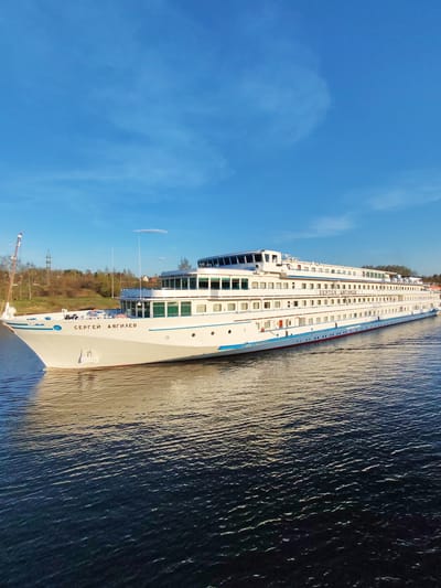 A large white cruise ship named "Сергей Дягилев" is sailing on a body of water. The ship is multi-decked and has numerous windows. The water is dark, with light reflecting on its surface. The sky is blue with some clouds. A small amount of vegetation, possibly trees, is visible on the far shore. Based on the presence of the ship and possible location details, this could be near the city of Базарово, Russia, but the surroundings are somewhat indeterminate from the image itself.