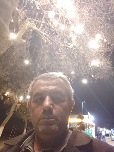 A middle-aged man with short, graying hair and stubble stands in front of a tree decorated with numerous small, warm-toned lights. In the background, there is a brightly lit area that appears to be a marketplace or shopping center with people walking around. A tall light pole is visible. The scene appears to be at night, located somewhere in Sabir Rakhimov, Uzbekistan.
