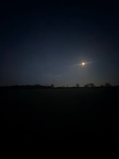 This media file captures a nocturnal landscape scene in Hœrdt, France, dominated by the brilliant light of the moon. The sky occupies the upper portion of the image, presenting a gradient of dark blue-grey that deepens to black away from the light source.

The primary subject is a strikingly bright, yellow-orange moon, positioned centrally in the upper half of the frame. It appears as a small, intense sphere, emitting a strong, almost vertical beam or lens flare that cuts through the atmospheric haze, illuminating the surrounding sky with a soft glow.

Below the illuminated sky, a faint, dark silhouette of a distant tree line or vegetation marks the horizon, serving as a subtle separation from the foreground. The ground in the lower portion of the image is extremely dark, almost black, suggesting an unlit field or open rural land.

The image conveys a sense of peaceful solitude characteristic of a clear, moonlit night. There are no people or specific objects discernible beyond the natural elements. No activity is taking place; it is a static landscape shot. The time of day is clearly night, and the weather appears calm, allowing the moon to shine brightly, possibly through some atmospheric mist or clouds contributing to the diffused light effect. There is no visible text.
