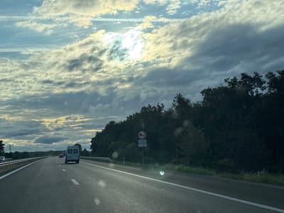 The image depicts a highway scene under a cloudy sky with the sun breaking through the clouds. The road is paved and marked with white lines. In the foreground, a gray van is visible in the right lane, driving away from the camera. To the left, another vehicle is partially visible on the highway. On the right side of the road, a dense line of trees and bushes forms a natural barrier. A road sign with circular symbols and text indicating traffic regulations is placed along the roadside. The overall environment suggests a rural or semi-rural highway setting.