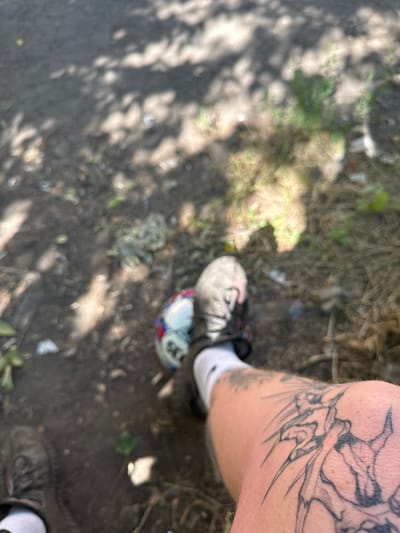 The image depicts a close-up view of a person's leg with a tattoo, a soccer ball, and footwear. The leg, which is tattooed with what appears to be a mythical creature or an abstract design, is in the foreground. A soccer ball, partially visible, is positioned near the person's foot, which is clad in a black and gray athletic shoe with white socks. The background is out of focus and appears to be an outdoor environment, possibly a park or a dusty area, with shadows from trees cast on the ground. The lighting suggests it is daytime.