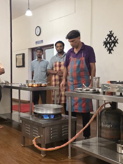 In Perumannur, India, three men are gathered around cooking stations in what appears to be a culinary class or demonstration. The man in the foreground, wearing a purple shirt and a checkered apron, stands by a gas cylinder and a stainless steel cooking unit where a large pot is heating on a lit blue flame. To his left, two other men, dressed in light blue shirts and aprons, observe or participate. One of them is wearing a checkered apron and is positioned behind a stainless steel table laden with copper pots. The background reveals a classroom-like setting with a clock on the wall, a framed artwork, and fluorescent lighting overhead. The floor is made of polished wood.