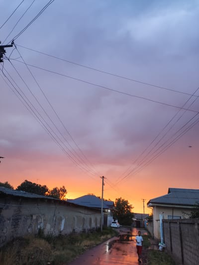 In the city of Mingchinar (Blagoveshchensky), Uzbekistan, a young boy in a white t-shirt and red shorts walks down a wet, unpaved road reflecting the vibrant colors of a sunset. The sky above is a dramatic gradient of purple, pink, and orange, with scattered clouds. Overhead, a network of power lines crisscrosses the frame, supported by utility poles. To the left, a long, weathered wall with visible cracks separates a property from the road. Various houses with tiled roofs are visible on either side of the street, some with fences. A white car is parked on the left side of the road, further down. The overall scene depicts a moment of atmospheric beauty in a residential area of the city.