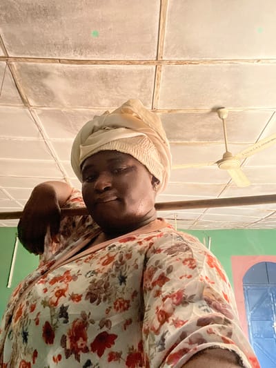 An individual, identified as female, is depicted in an indoor setting within Makera, Nigeria. She has a dark complexion and wears a light beige headwrap with a shirred band over her forehead. Her attire consists of a light-colored garment patterned with red, brown, and dark floral motifs, featuring a brown collar. She is positioned with her torso angled, and her head turned towards the camera, exhibiting a slight smile. A portion of her left arm is visible, bent at the elbow, and a small earring is present in her visible right ear.

In the background, a grid-paneled ceiling with wooden or metal strips is visible. A cream-colored three-bladed ceiling fan is mounted in the upper right quadrant of the frame; text partially visible on one of its blades reads "ESO OF C.P.S. KAKURI BAH...". The walls include a section painted green on the left (viewer's right) and a light pink/peach section further right, behind which a dark blue arched door or archway is partially visible. The scene is illuminated by ambient light.