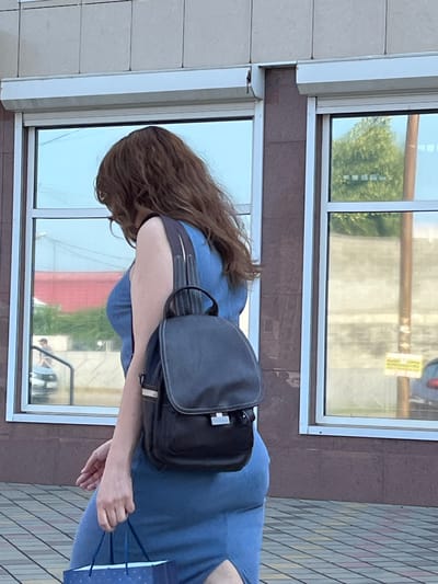 A woman with long, wavy brown hair is walking away from the camera, from left to right. She is wearing a sleeveless blue dress and a black backpack with a flap closure and silver buckles. Her left hand is carrying a blue gift bag with white polka dots. She is walking on a tiled sidewalk in front of a building with large, reflective windows. The windows reflect trees, buildings, and a car. A roller shutter is visible above the windows. The scene appears to be in Apsheronsk, Russia, based on the provided context.