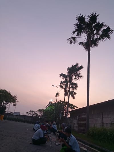 A low-angle shot depicting an outdoor scene at dusk in Nglundo, Indonesia. A group of about 15 individuals, mostly dressed in light-colored shirts and some wearing head coverings, are seated on the pavement along a curb. Some appear to be looking at their phones or interacting with objects in their laps. Empty cups and bottles are scattered near them. Behind the group, a tall gray fence borders the area, and some green grass is growing along the base of the fence. Several palm trees of varying heights stand tall to the right, silhouetted against the pink and purple sky. A street lamp is seen between them and the sign "NO SMOKING ZONE" can be seen written on a board on the opposite side of the road, further away from the scene. The pavement is made of gray brick or stone.