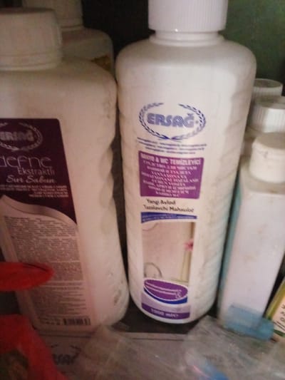 The image shows a close-up view of several bottles of cleaning products on a shelf. The bottles are white with labels in various colors. One bottle on the left has a dark purple label with text in white. The bottle in the center is also white with a purple and blue label. Text on this label is in Turkish and includes the brand name "ERSAĞ". The bottle on the right is white with a white cap and no visible label. The background is dark and out of focus. The scene appears to be indoors, possibly a home or a shop. There are no people visible in the image. The location of Kara-Bak, Kyrgyzstan is not discernible from the image itself.