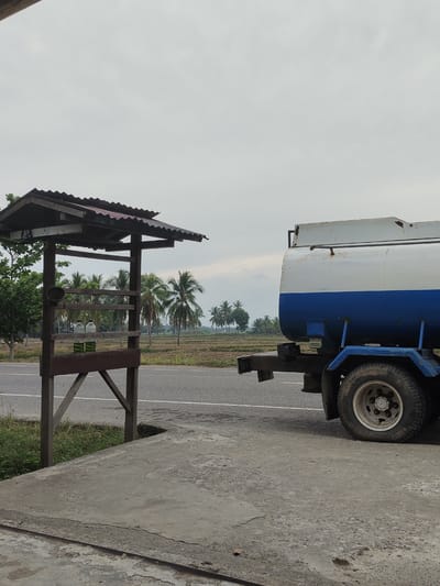 A blue and white water truck is parked on the side of a road in Aluekeujruen, Indonesia. Next to the truck is a simple wooden structure with a corrugated metal roof. The structure appears to be a roadside stall or shelter. Two plastic bottles filled with green liquid are visible inside the structure. The background features a field with scattered palm trees under an overcast sky. The ground in the foreground is a concrete surface.