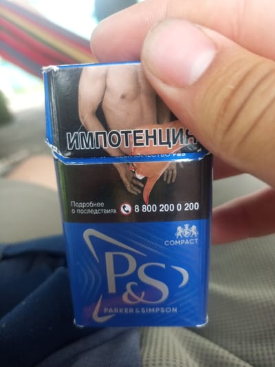 A hand is holding a blue cigarette pack that is part of a warning about impotency. The pack has a close-up image of a person's torso, with text in Russian that translates to "impotency." Below this, there is text that reads "Learn more about the consequences" and a phone number: "8 800 200 0 200." The pack also features the P&S logo with "COMPACT" and "PARKER & SIMPSON" written below it. The background suggests an outdoor setting with a hammock visible. This image was taken in Kalach, Russia.