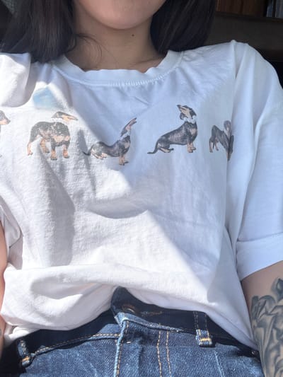 The image shows a person wearing a white t-shirt with dachshund dog prints, and blue denim jeans. Part of the person's torso, shoulder, upper arm with a tattoo, and dark hair are visible. The background is blurred and indistinct. There is no indication of Meshtitsa, Bulgaria in the image.