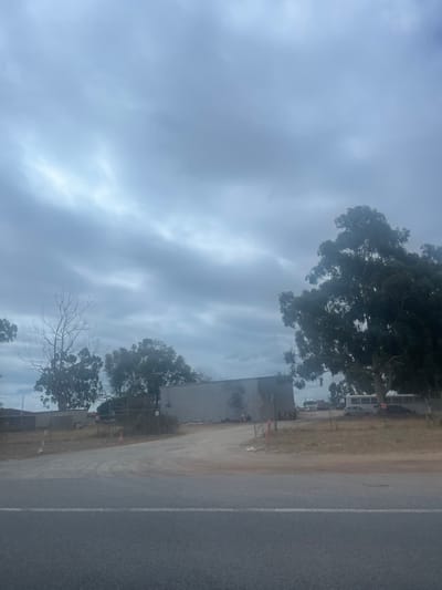 A cloudy sky dominates the scene. A grey building with the number "1987" visible is positioned to the right. Trees frame the building, some in full leaf and others bare. A road with a white dividing line is in the foreground. The ground to the side of the building appears dry and dusty. The overall impression is one of a bleak, overcast day with a hint of construction or industrial activity.