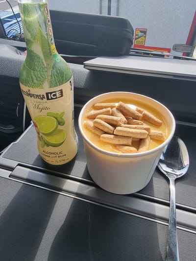 The image depicts a close-up view of a small table or tray, likely inside a vehicle, with food and a drink. In the foreground on the left is a green bottle labeled "LA RECOMPENSA ICE SABOR Mojito" with an image of limes and mint. The bottle states "ALCOHOLIC DRINK WITH RUM". To the right of the bottle is a white disposable cup filled with a yellow, creamy substance, topped with several small, light brown cylindrical crackers or biscuit pieces. A silver spoon rests to the right of the cup. In the background, a black bag is visible, and a sticker with red and yellow markings reading "ATTENTION ANGLES MORTS" is affixed to a reflective surface. The overall setting appears to be a casual, possibly on-the-go meal. The location of Portsmouth, United Kingdom, is not visually identifiable from the image content.