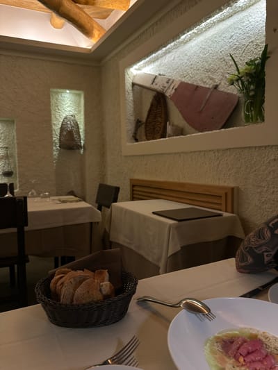 The image captures an interior dining scene, likely at a restaurant, in Agropoli, Italy. The camera's perspective is slightly angled. A table setting occupies the foreground with a white plate containing a dish of raw fish, silverware including a spoon and fork, and a bread basket filled with sliced bread. Seated at the table is a person whose tattooed leg is visible. The table is covered with a white tablecloth. In the background, the restaurant's decor is visible: textured white walls with inset display niches featuring decorative objects. A niche near the camera includes greenery and an object with a pink tone. The restaurant appears dimly lit, with light sources focused on the decorative features and tables.