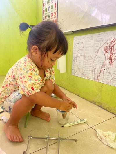 A young barefoot child, identified as female, is depicted squatting on a tiled floor in Desa Batu Layar, Indonesia. She wears a light-colored floral blouse with yellow, red, and blue patterns, and light-colored patterned shorts. Her dark hair is pulled into a single ponytail with a green elastic, and she wears small hoop earrings. Both hands are engaged with a clear plastic cup containing soil and a brown, organic object. Her hands are visibly soiled. On the floor nearby are a white dry-erase marker with a black cap, a green pen, a crumpled piece of white tissue with dirt smudges, and a clear acrylic structure with metal screws and wing nuts. The background features a vibrant lime-green wall with a white dry-erase board mounted on it. The whiteboard contains handwritten lists of "Colours," "Fruits," "Countries," "Numbers," and "Family," partially obscured by extensive red scribbles. Above the whiteboard, a portion of an alphabet chart (A-Z) is visible on the green wall. The child is focused downwards, intently observing or manipulating the contents of the cup.