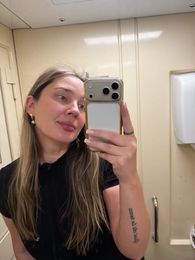 A woman is taking a selfie in what appears to be a bathroom. She is holding a smartphone with a gold case and a three-lens camera. Her right arm is visible, revealing a tattoo. She has long, highlighted hair, light skin, and is wearing earrings. The background consists of a light-colored wall with horizontal paneling, a toilet paper dispenser, and a metal handle. The location is unknown as there is no city of Пугачёво in Russia.