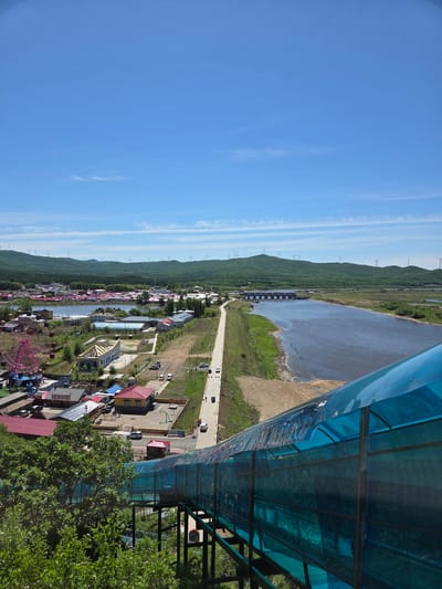 This high-angle, panoramic shot, taken from an elevated viewpoint in Jiejinkou, China, showcases a vibrant rural landscape under a clear blue sky. The image is dominated by natural and man-made features stretching across the scene.

In the immediate foreground, a striking blue, transparent canopy structure extends across the upper right portion of the frame, supported by metal beams. This structure suggests a covered walkway or viewing platform, from which the photograph was likely taken. Lush green foliage, including tree leaves, is visible in the bottom right, hinting at a natural setting around the viewpoint.

Looking down, the scene unfolds to reveal a small settlement or town situated along the left side. It features numerous buildings with varied architectural styles, some sporting brightly colored roofs in red and blue hues. A prominent pink Ferris wheel stands out, indicating the presence of an amusement or entertainment area within the town. A road, with a few cars parked or in motion, weaves through the settlement.

To the right of the town, a wide body of water, possibly a river or a reservoir, meanders through the landscape. Its banks are a mix of sandy stretches and green vegetation. A dam or a series of sluice gates is visible spanning part of the water body, suggesting water management. Another road runs parallel to the water's edge.

In the mid-ground and background, rolling hills or mountains, densely covered in green trees, define the horizon. A distinctive feature of these hills are numerous tall, white wind turbines, standing majestically on the ridgelines, indicating a source of renewable energy. The sky above is a brilliant blue with only a few wispy white clouds, suggesting clear and pleasant weather. The overall impression is one of a peaceful, well-developed rural area, possibly a tourist destination, enjoying a bright, sunny day. No discernible people or visible text are present in the image.