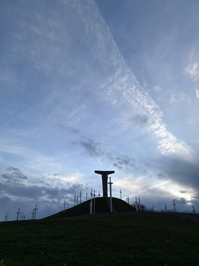 The image depicts a low-angle view of the Memorial of Georgian Soldiers in Zemo Nichbisi, Georgia. The sky is predominantly light blue with streaks of white clouds and a distinct contrail. A green hill rises from the lower right, dotted with numerous white sword-shaped monuments. At the crest of the hill, a large, dark sword-shaped monument stands out against the sky.