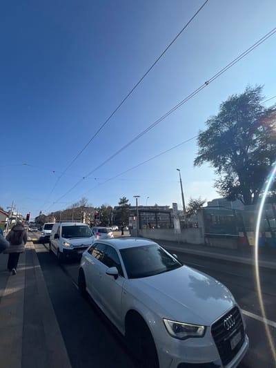 This image, taken in Meyrin, Switzerland, captures a bright, sunny daytime street scene from a low angle, looking upwards and along a road. The clear, deep blue sky occupies a significant portion of the upper frame, marked by visible lens flares from the sun.

Prominently featured on the right side is a white Audi sedan, its front grille and headlights distinctly visible as it faces away from the viewer. Behind it, a white van and several other cars are seen, suggesting a line of traffic or parked vehicles. Further down the street, red traffic lights are discernible. The lower right corner reveals a partial view of a person's legs and shoes, indicating a pedestrian's viewpoint near the roadside.

Numerous thin, dark overhead power lines, likely catenary lines for a tram or trolleybus, stretch across the sky from the left to the right side of the image, with small insulators and connection points visible. The background consists of urban buildings and structures along the street, interspersed with trees. A tall street light pole stands upright in the mid-ground. The overall impression is a vibrant, active street on a clear weather day. No visible text is clearly legible.