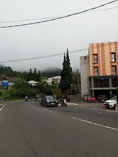 The image captures a foggy mountain road scene within the city of Phoxay, Laos. The sky is uniformly overcast and grey, with a dense layer of fog or low clouds obscuring the peaks of thickly forested mountains in the background.

The foreground features an asphalt road that curves to the left, delineated by a solid white line on its right margin. On the right side of the road, a multi-story building stands. Its upper section displays alternating vertical stripes of beige and orange, punctuated by dark-framed rectangular windows. The ground floor of this building features a dark grey facade with large glass panels, and an adjoining section to its left exhibits a light grey, textured block pattern. A paved area in front of the building serves as a parking space, containing a red compact car, a dark SUV, and a white SUV positioned near an orange traffic cone.

Two prominent, tall, slender conifer-like trees with dark green foliage are situated between the road and the building on the right.

Active traffic is present on the road. A dark grey compact multi-purpose vehicle (MPV) bearing license plate '7021 MUN' is moving towards the viewer. Alongside it, to the right, a person wearing a helmet operates a white scooter. Further down the curving road, another individual on a scooter is visible behind the MPV, with at least two additional scooters and riders discernible in the distance on the left side of the curve. All visible individuals operating scooters are wearing helmets.

To the left of the road, on a verdant hillside, several white-painted residential structures are nestled among abundant green trees and foliage. A partial blue rectangular sign mounted on poles is visible on the far left. Multiple dark utility wires traverse horizontally across the upper frame of the image.