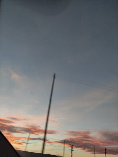 The image displays a sky at either sunrise or sunset, viewed from what appears to be a moving vehicle. The sky is predominantly split diagonally; the upper left portion features a muted blue-grey color, while the lower right is bathed in the warm hues of orange and pink, indicative of the sun's low position.
A tall, slender utility pole or mast stands slightly to the left of the center, exhibiting a subtle blur that suggests the photo was taken from a moving perspective. Several thin power lines stretch horizontally across the sky, connected to additional utility poles that are visible on the far right edge of the frame. A dark, indistinct shape in the bottom-right corner likely represents a part of the vehicle's interior, framing the natural spectacle.
The weather appears clear with scattered clouds, emphasizing the dramatic sky colors. There is no visible text within the image. The presence of utility infrastructure suggests an industrial or semi-rural setting within Torre Pacheco, Spain.