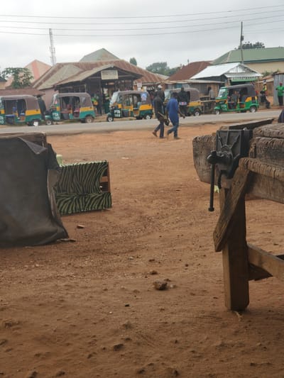 In Barikin Sabon Gidan Kanat, Nigeria, an outdoor scene unfolds with a row of auto-rickshaws, commonly known as Keke Napeps, parked on the side of a dirt road. The vehicles, primarily yellow and green, bear markings such as "PTA 1005" and "PTA 1389". Several individuals are present, with two men walking in the foreground, one in a blue shirt and the other in dark clothing. Behind them, a few more people are visible near the Keke Napeps and what appears to be a market stall with a sign that reads "Newborth Stores". The environment is dusty, with a wooden workbench and vice prominently featured in the right foreground. A black tarp covers an object to the left, and a seat covered in a zebra-patterned fabric is also visible on the ground. The sky is overcast, suggesting a cloudy day. Buildings with corrugated iron roofs are situated in the background.