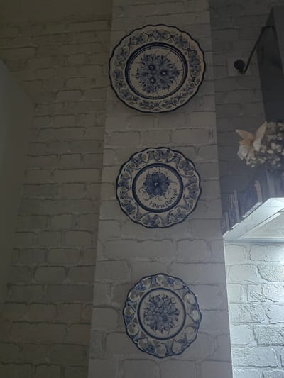 Three decorative ceramic plates with blue floral patterns are mounted on a textured white brick wall. The plates are arranged vertically, with the topmost plate being the largest and the bottom two being smaller and identical. The middle plate has the inscription "Fátima Portugal" on it. To the right of the plates, part of a silver kitchen appliance or ventilation system is visible, along with a shelf containing books and a decorative dried floral arrangement. The scene is indoors, likely in a kitchen or dining area, with soft lighting. The location in Offenbach am Main, Germany, is not discernible from the image itself.