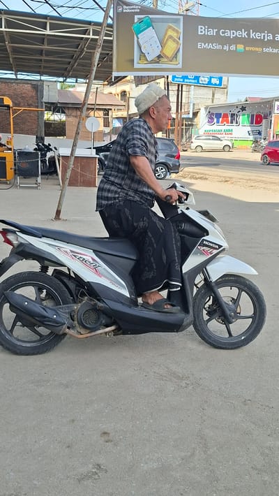 A middle-aged man wearing a plaid shirt, black patterned pants, and a white cap is riding a white and black scooter. He is positioned in the middle of the frame, slightly angled to the right. The man is looking to the right, and his hands are on the scooter's handlebars. The scooter is parked on a paved surface, likely a road or parking lot. In the background, there are several buildings, including what appears to be a commercial establishment with a sign that reads "STAR KID." There are also other vehicles visible, including a dark-colored car and a red car. The sky is clear and blue. The overall impression is an outdoor scene in an urban or semi-urban environment in Blangpeuria, Indonesia.