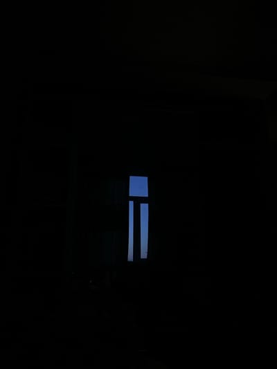This image depicts an interior space in Navoiy City, Uzbekistan, characterized by profound darkness. A rectangular window is positioned on the right half of the frame, serving as the sole source of illumination. Through this window, a clear, solid blue sky is visible, suggesting daylight conditions outside. The window structure includes two distinct vertical dark elements, consistent with window frames or mullions, dividing the visible sky into three vertical sections. The surrounding interior walls, floor, and ceiling are entirely obscured by deep shadow, rendering any additional subjects or objects undetectable. No individuals or specific actions are discernible within the scene.