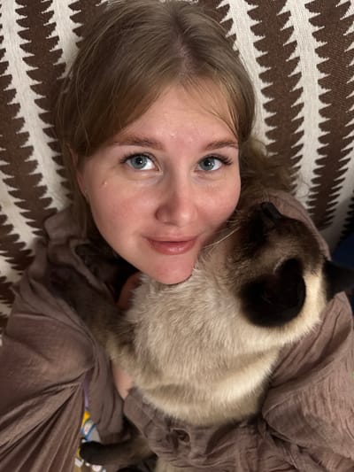 The image is a close-up, overhead shot of a young Caucasian woman with light brown hair, blue eyes, and fair skin, who is lying down and holding a Siamese cat. The woman is wearing a taupe-colored garment. The cat is light-colored with dark points on its face and ears. They are positioned on a patterned surface, likely a couch or bed, with a brown and white geometric design. The environment appears to be indoors. There is no indication of the city of Буб, Russia in the image itself.